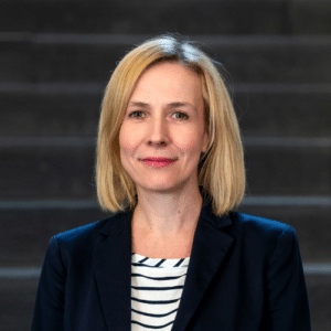 Porträt einer Frau mit blonden Haaren, in einem Blazer, vor einer dunklen Treppe.
